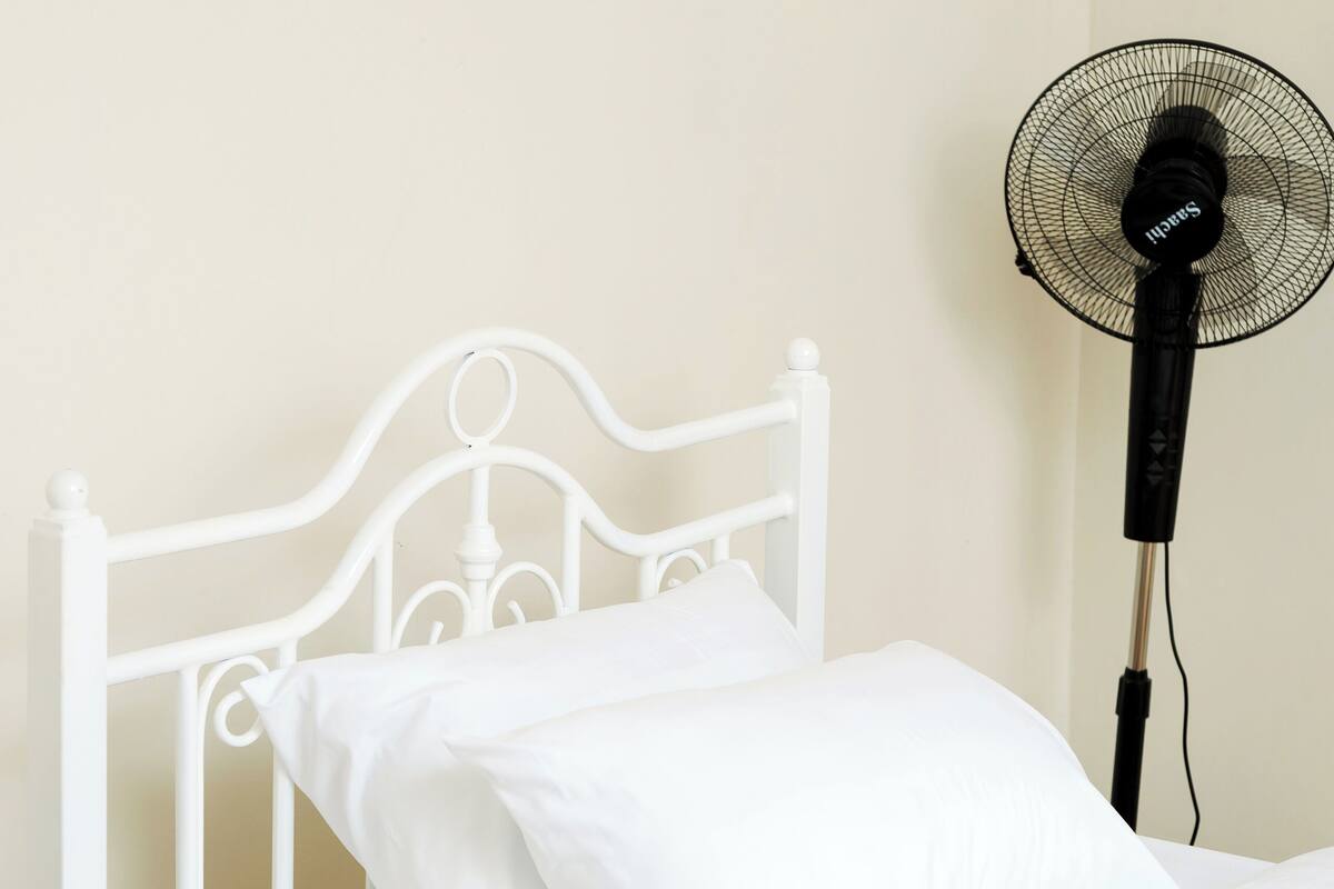 Una cama metálica blanca con colchón y almohadas blancos, una mesita de noche y un ventilador de pie.