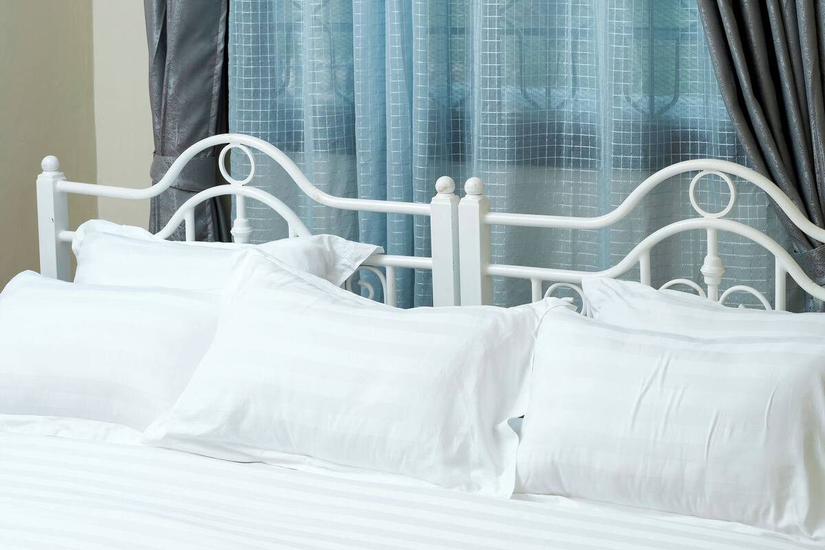 Una cama con ropa blanca, estructura metálica, cortinas y ventana con vistas.