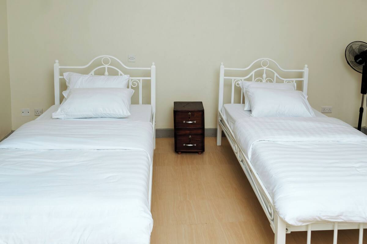 Una habitación con dos camas individuales, una mesita de noche de madera y un ventilador de pie.