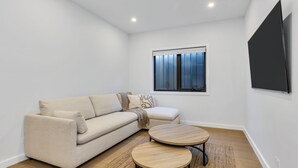 A modern living room with a beige sofa, a round wooden coffee table, and a large window with blinds. - The Base at Abbott. Unwind with Private Spa -14 Abbott Street (Jindabyne)