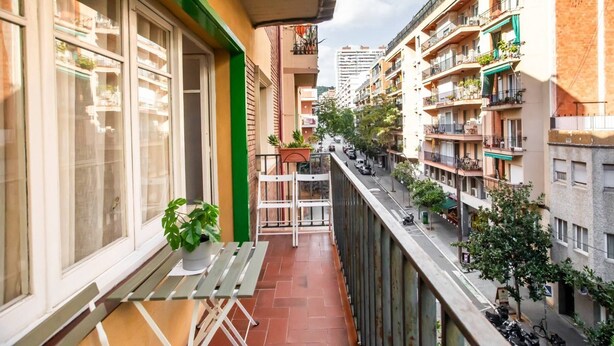 Un balcon étroit avec une table et des chaises, un pot de fleurs et une vue sur une rue animée, bordée d’immeubles et de voitures garées.
