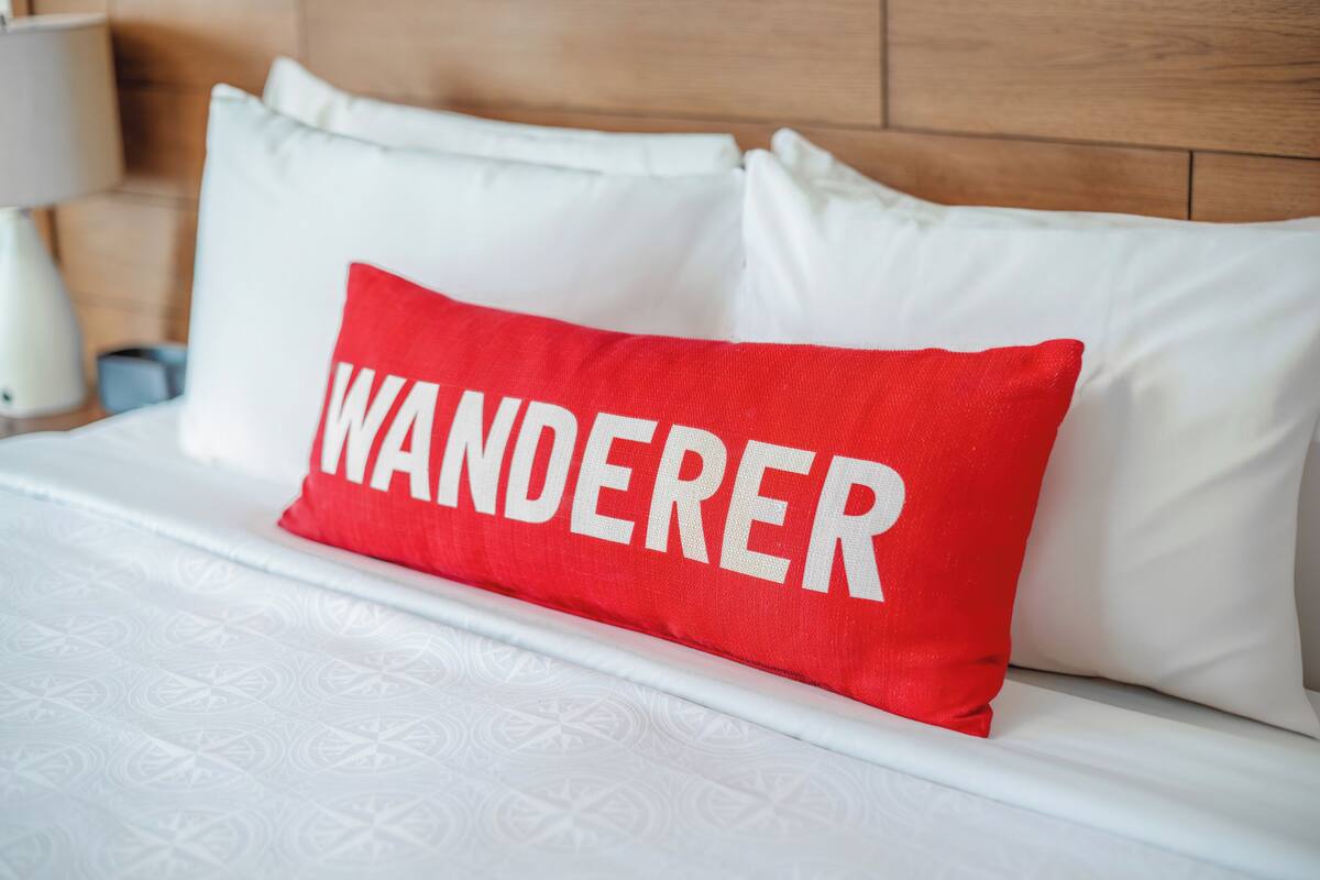 A bed with white bedding and a red pillow with the word "WANDERER" on it.