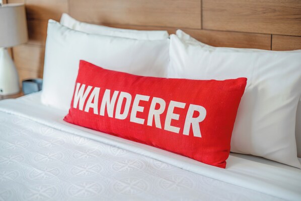 A bed with white bedding and a red pillow with the word "WANDERER" on it. - Compass Hotel Beaufort by Margaritaville (Beaufort)