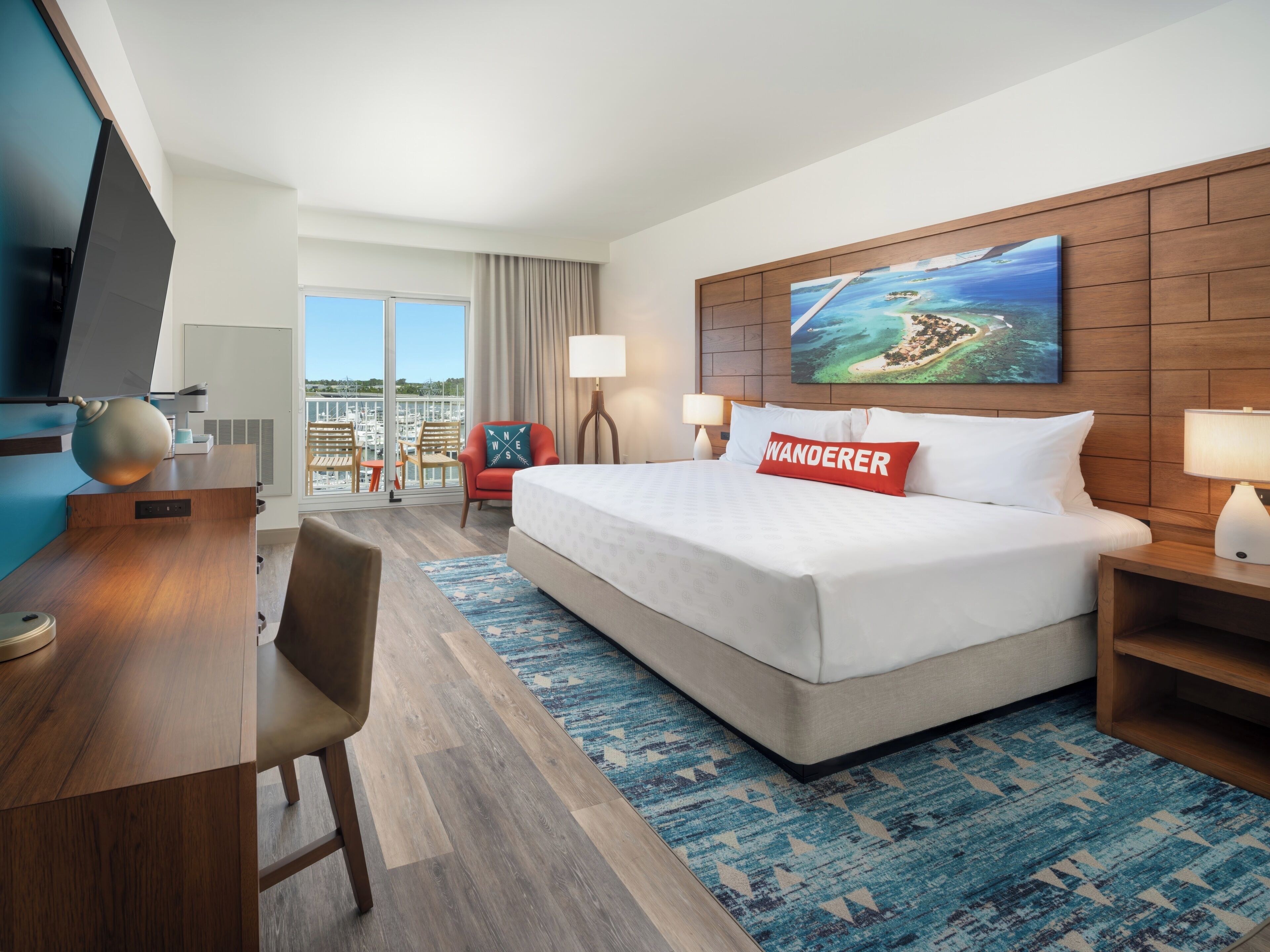 Une chambre d’hôtel avec un grand lit, un bureau avec une télévision, une chaise, une fenêtre donnant sur le port de plaisance et un tableau au mur.