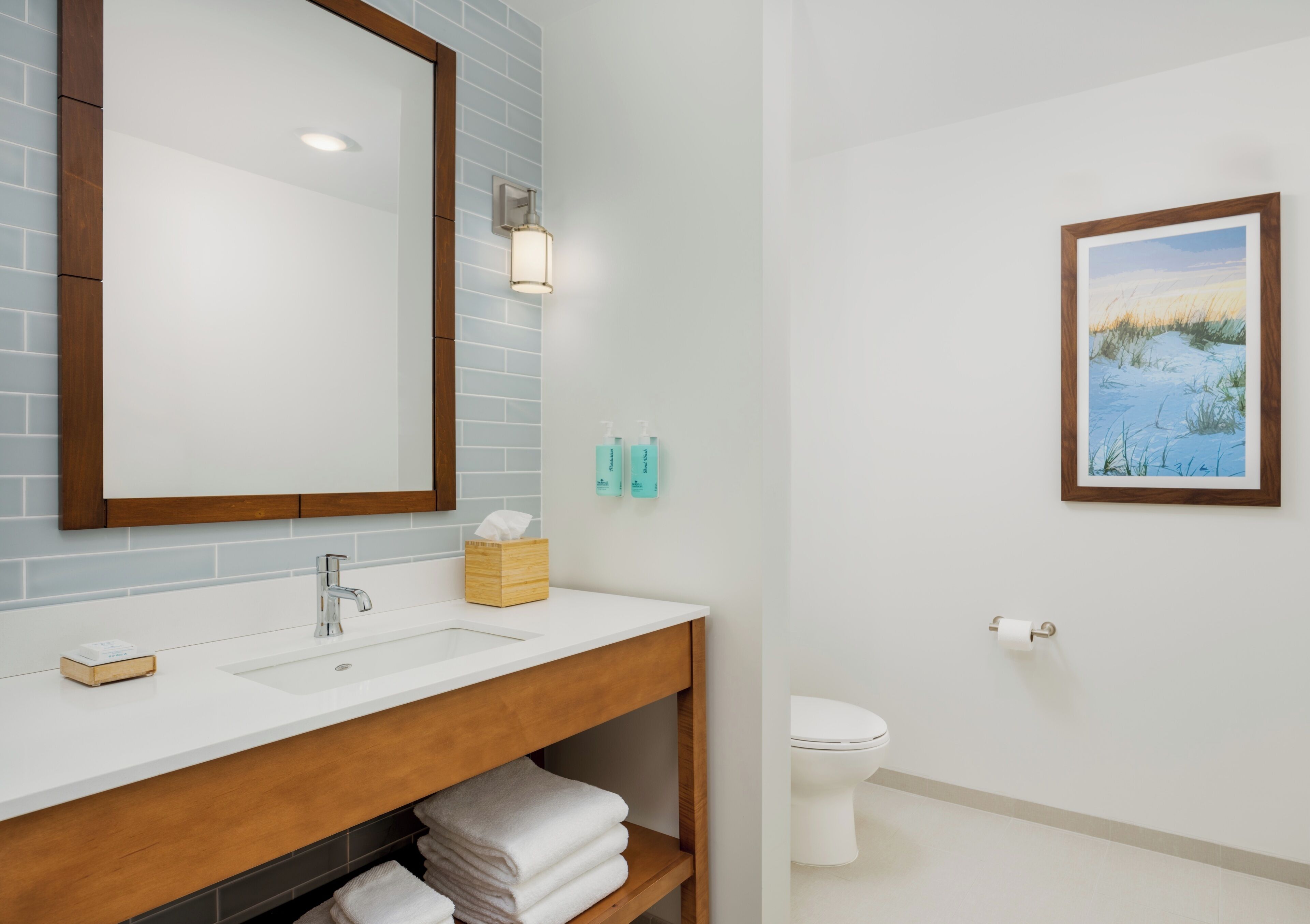 A bathroom with a white sink, a mirror, a wooden vanity, a toilet, and a framed picture on the wall.