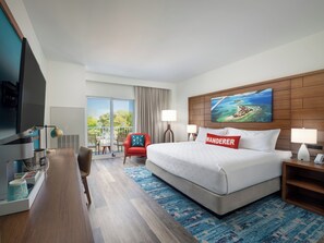 A hotel room with a large bed, a desk with a computer, a TV, a red armchair, and a balcony with a view of greenery. - Compass by Margaritaville Beaufort (Beaufort)