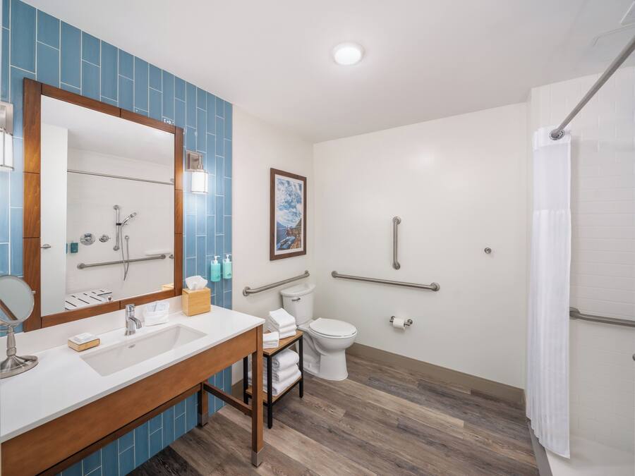 Un baño moderno con tocador de doble lavabo, inodoro con barra de apoyo, zona de ducha y dispensador de papel higiénico blanco.