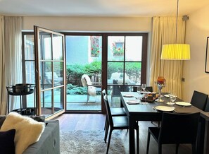 A dining area with a table set for two, a sofa, a lamp, and a view through a glass door to an outdoor area with plants.