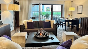 A modern living room with a sofa set, a coffee table with a tray of glasses, a dining area with a table and chairs, and a large window with a view of greenery. - Exclusive deluxe suite in the heart of Kitzbühel (Kitzbühel)