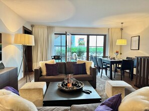 A modern living room with a sofa set, a coffee table with a tray of glasses, a dining area with a table and chairs, and a large window with a view of greenery.