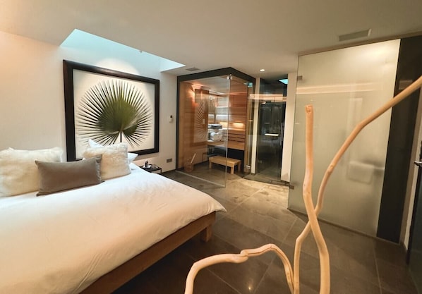 A modern bedroom with a large bed, a framed artwork, a wooden bench, and a glass door leading to a bathroom.