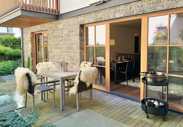 A wooden deck with a table and chairs, a sliding glass door, and a metal cart.