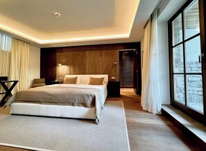 A modern bedroom with a large bed, a bedside table, a lamp, and a view of a stone wall through a window.
