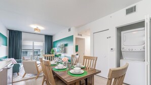 A modern, well-lit dining area with a wooden table set for four, surrounded by wooden chairs. A kitchen with a microwave and a refrigerator is visible to the right.