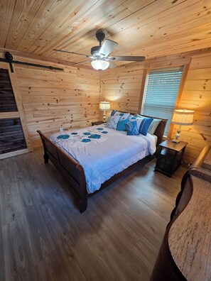 Quarto com teto de madeira, cama com colcha florida, mesa de cabeceira com abajur e janela com persianas.