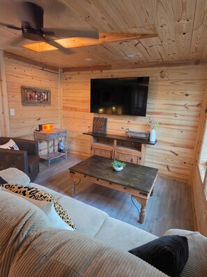 A cozy room with a wooden interior, a flat-screen TV mounted on the wall, a small table with a plant, and a sofa with patterned cushions.