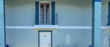 A two-story building with a balcony, a door, and two wicker chairs outside.