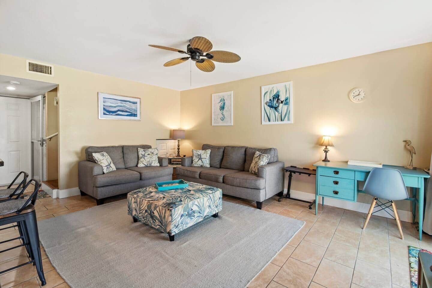 Ein Wohnzimmer mit grauem Sofa, Hocker mit Muster, Schreibtisch und Stuhl sowie Deckenventilator.