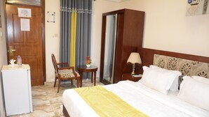 A hotel room with a bed, a desk, a chair, a small table with a lamp, and a refrigerator. - Hunters Royal Apartments (Madina)
