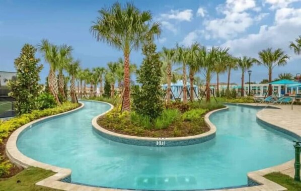 A curved swimming pool surrounded by palm trees and greenery, with a clear sky.