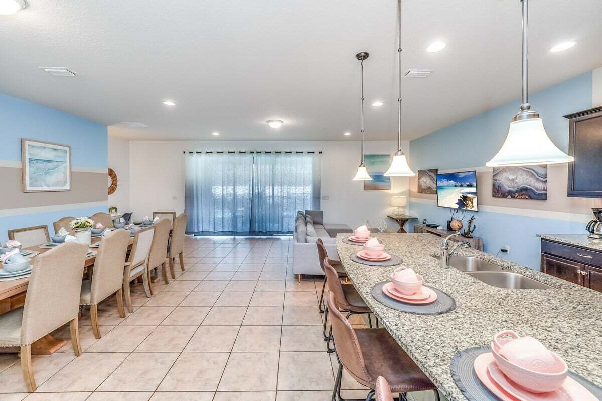 A modern kitchen with granite countertops, a dining area with a long table, and a large window with curtains.