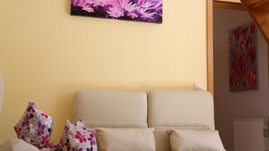 A room with a beige sofa, floral-patterned pillows, and a wall-mounted painting of purple flowers. - Ferienwohnung in Freital, Dresden (Freital)