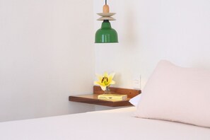 A bed with a white pillow, a bedside table with a green lamp, a yellow flower, and a book.
