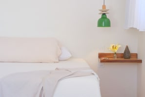 A bed with a white blanket and pillows, a green hanging lamp, and a wooden shelf with a vase and a flower.