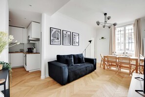Ein modernes Wohnzimmer mit einer schwarzen Couch, einem Holzespeisesaal und Stühlen. Es gibt eine Küchenzeile mit weißen Schränken und einem Fischgrätenboden.