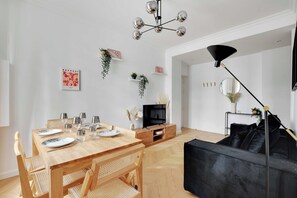 Ein modernes Wohnzimmer mit einem Holzesstisch, gedeckt für sechs Personen, einem schwarzen Sofa und einem Fernseher auf einem Holzgestell.
