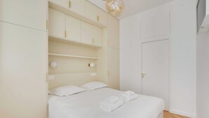 A hotel room with a bed, white walls, a wardrobe with multiple shelves, and a door. - Spacious Cosy Apartment -2br/8p- Pl. de la Nation (Paris)