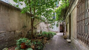 A narrow alley with a concrete wall on one side and a building with barred windows on the other. There are potted plants and ferns along the wall, and the ground is covered with fallen leaves. - Cosy Apart - 3br/6p - Jardin du Luxembourg/odeon (Paris)