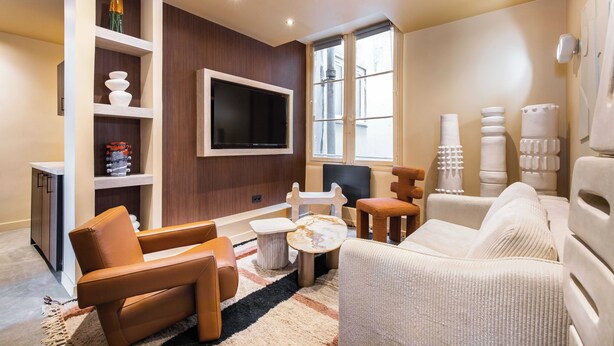 A modern living room with a flat-screen TV mounted on the wall, a brown leather sofa, a white armchair, and a small round coffee table. - Gorgeous Apartment - 2br/8p - Le Marais (Paris)