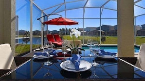 Un comedor con mesa de vidrio, paraguas rojo y vistas a una piscina y casas.