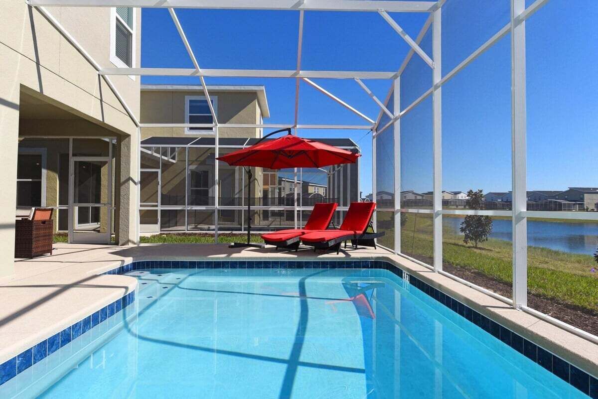 Una piscina con sillas reclinables y una sombrilla roja, rodeada por un cerramiento de cristal con vistas a un lago y edificios.