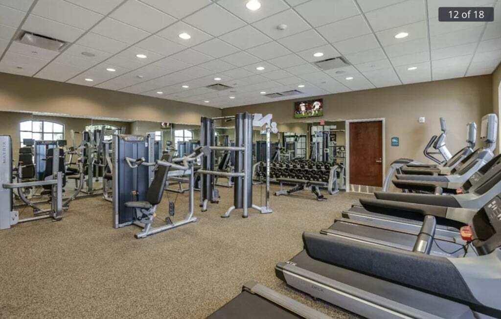 A gym with various exercise equipment, including treadmills and weight machines, arranged in a spacious room with a tiled ceiling and large windows.