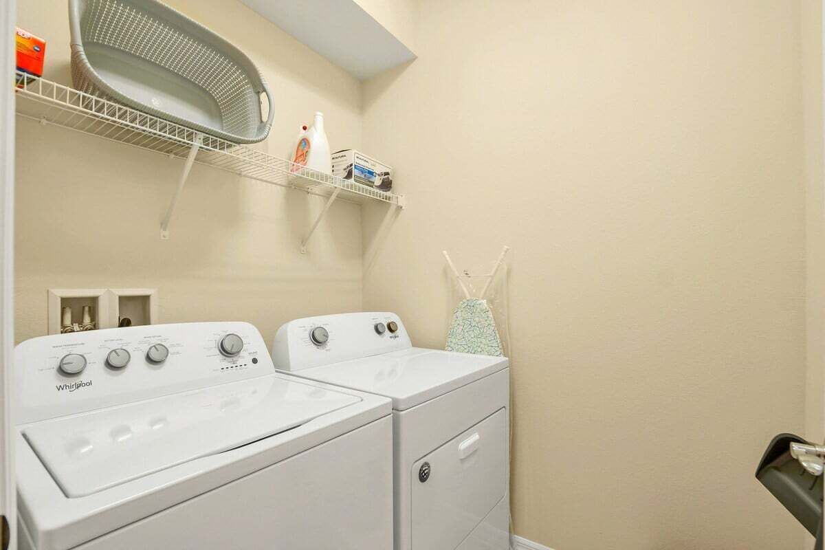 Une buanderie équipée de deux lave-linge et deux sèche-linge Whirlpool blancs, d’une étagère métallique au-dessus pour ranger les produits de nettoyage, et d’une petite fenêtre avec un rideau.