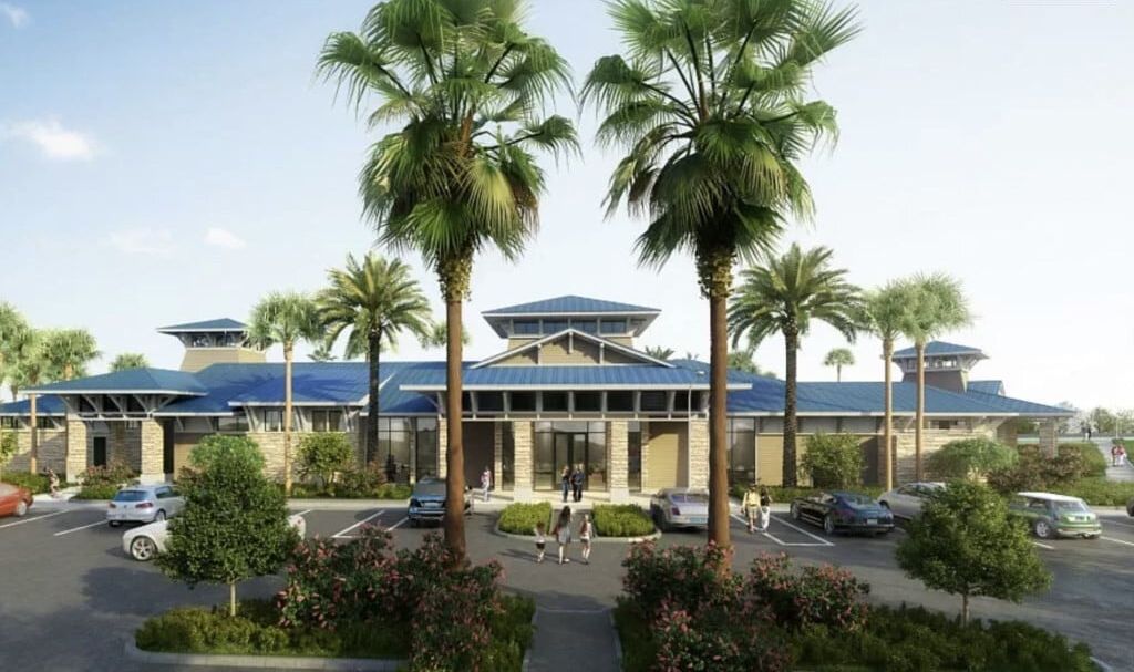 A hotel with a modern architectural design, featuring a central entrance flanked by palm trees, a parking lot, and a clear sky.