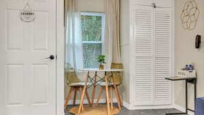 A room with a white door, a window with blinds, a small table with a plant, a chair, a rug, and a closet with a 'HOME' sign.