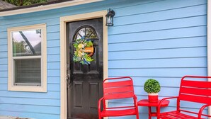 A blue house with a black door, a window with blinds, and two red chairs with a small table and a potted plant.