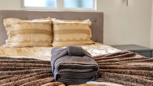 A neatly made bed with a folded towel in the foreground, a window with blinds, and a wall-mounted light fixture.
