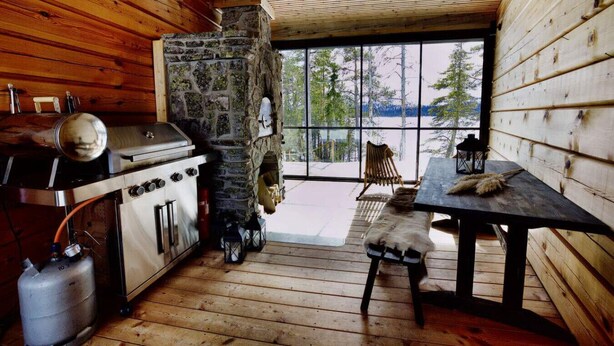 Interior de una cabaña con chimenea de piedra, paredes de madera, una mesa de comedor y vista a árboles a través de ventanas amplias.