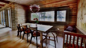 Interior de una cabaña de madera con una mesa de comedor, sillas y un ventanal que ofrece vistas a árboles.