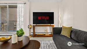 Un salon moderne avec un grand téléviseur à écran plat affichant le logo « NETFLIX », un canapé gris foncé, une table basse en bois et une plante en pot.