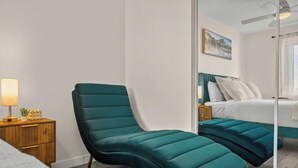Une chambre à coucher moderne avec un fauteuil recouvert d’un tissu turquoise, une table de chevet en bois avec une lampe et un lit avec une literie turquoise.