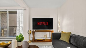 A modern living room with a large flat-screen TV displaying 'NETFLIX', a dark grey sofa, a wooden coffee table, and a potted plant. - Steps to 17 Ave, 2 queens, UG parking, AC (Calgary)