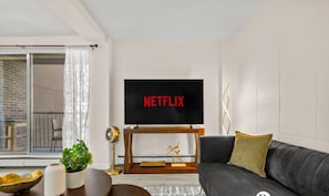 Een moderne woonkamer met een grote flatscreen-tv waarop 'NETFLIX' staat, een donkergrijze bank, een houten salontafel en een potplant.