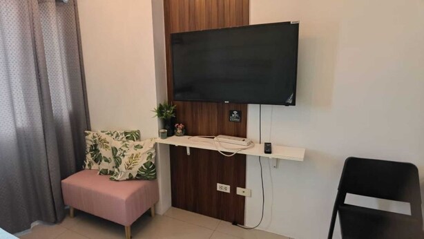 Habitación de hotel con una TV de pantalla plana montada en la pared, un pequeño estante de madera con una planta, un enchufe y una ventana con cortinas.