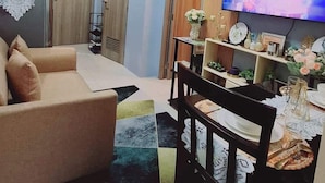 A living room with a sofa, a dining table with chairs, a TV mounted on the wall, and a ceiling light.