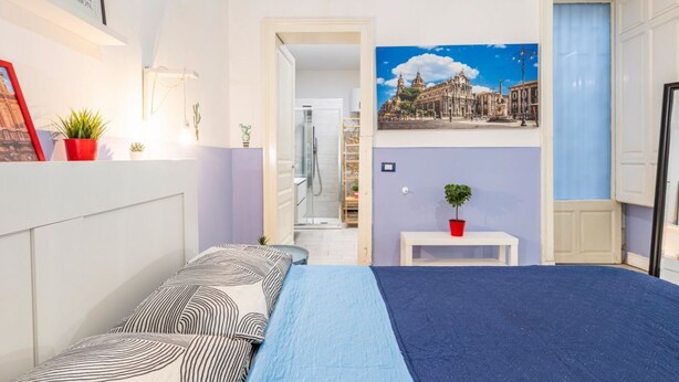 A bedroom with a bed, a painting, a potted plant, and a hallway leading to a bathroom. - GuestHost - Central apartment Caronda street (Catania)